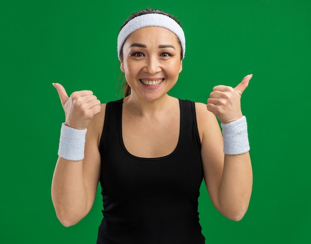 Молодая фитнес-женщина с повязкой на голову и нарукавными повязками с улыбкой на лице показывает палец вверх, стоя над зеленой стеной