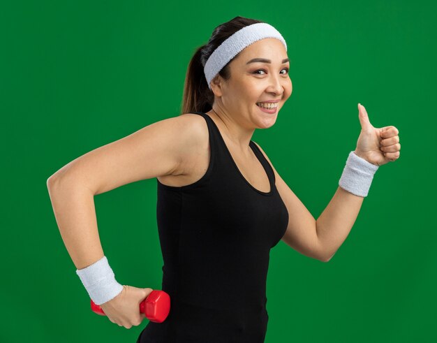 Молодая фитнес-женщина с повязкой на голову с гантелями делает упражнения, улыбаясь, показывает палец вверх, стоя над зеленой стеной