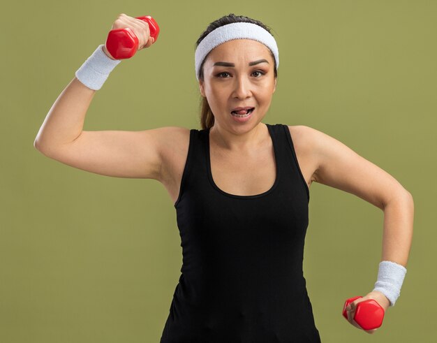 Young fitness woman with headband with dumbbells doing exercises strained and confident standing over green wall