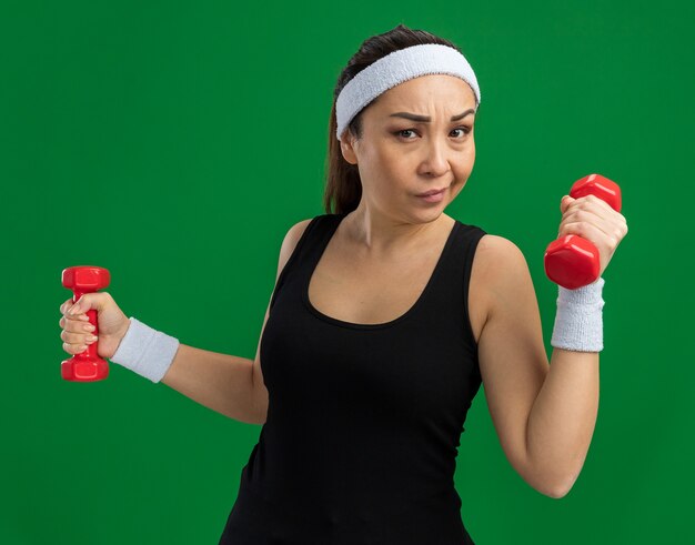 Young fitness woman with headband with dumbbells doing exercises strained and confident standing over green wall