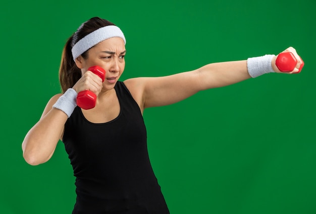 Young fitness woman with headband with dumbbells doing exercises strained and confident standing over green wall