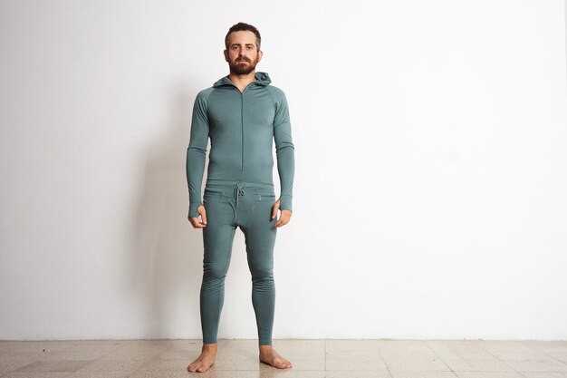 Young fitted bearded male wears thermal base layer suite , standing in front of white wall