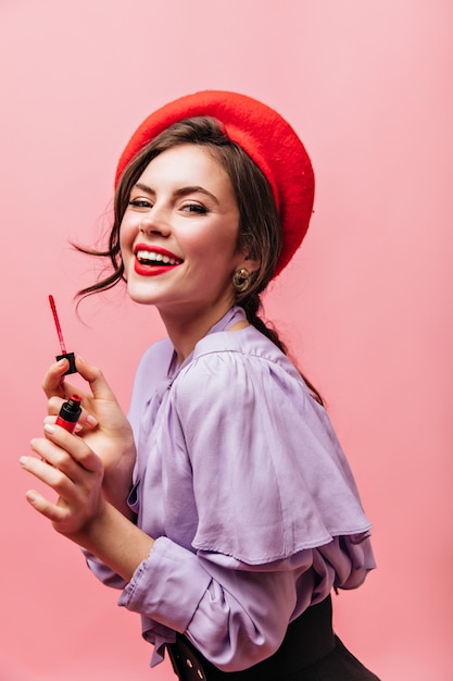 Foto gratuita la giovane donna civettuola dipinge le sue labbra con rossetto rosso. signora in camicetta viola e berretto luminoso in posa su sfondo rosa.