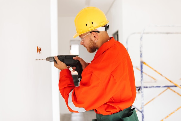 Young foreman in orange work clothes and yellow hardhat using electric drill at work in new apartments