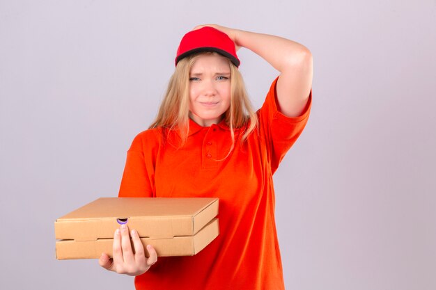 Young forgetful delivery woman in orange polo shirt and red cap holding pizza boxes holding hand on head as she realizes that she forgot about doing something important over isolated white back