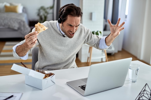 Young freelance worker feeling upset while eating and reading bad news over laptop at home