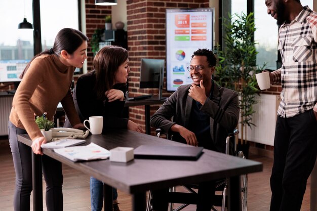 Young freelancer sitting in wheelchair and talking to business partners on work break, laughing and having fun in disability friendly office. Male worker with impairment chatting with colleagues.