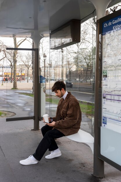 バスを待って、彼のスマートフォンを使用して駅で待っている若いフランス人男性