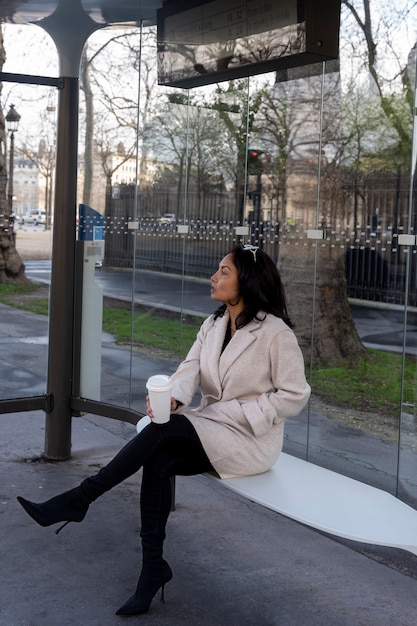 Young french woman waiting at the station for the bus and drinking coffee