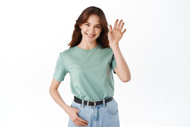 Бесплатное фото Молодая дружелюбная женщина машет рукой и здоровается, приветствует гостей, делает приветственный жест и широко улыбается, стоя на белом фоне
