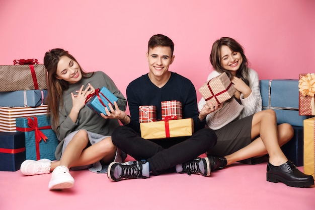 Young friends sitting among christmas gift boxes on pink