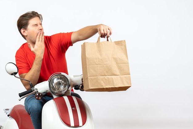 Young frowning courier guy in red uniform sitting on scooter giving paper bag on white wall