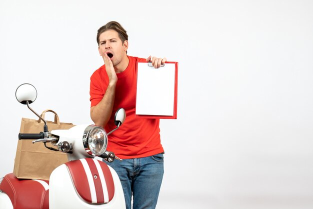 Young frowning delivery guy in red uniform standing near scooter showing document on white wall