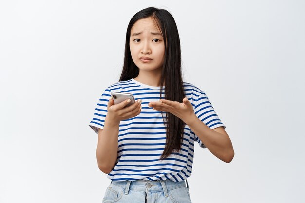 Young frustrated asian girl having problem with mobile phone, holding smartphone and pointing confused at screen on white