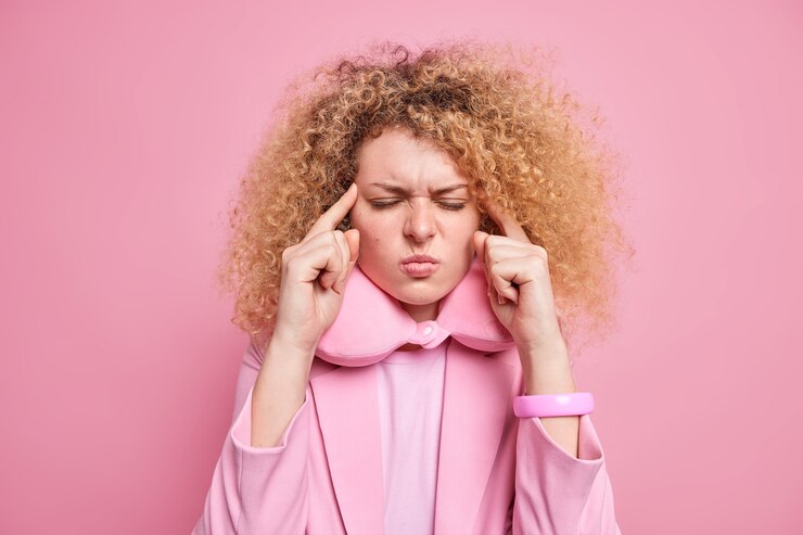 Young frustrated curly haired young woman keeps fingers on temples suffers unbearable headache prepares for sleep and having rest after hard busy day 