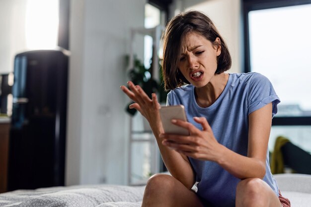 Young frustrated woman reading text message cell phone in disbelief while sitting on the bed at home