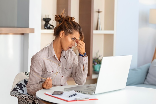 Молодая расстроенная женщина работая на столе офиса перед компьтер-книжкой страдая от хронических ежедневных головных болей