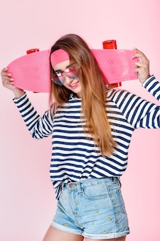 Young fun beautiful woman in glasses with skateboard on pink background