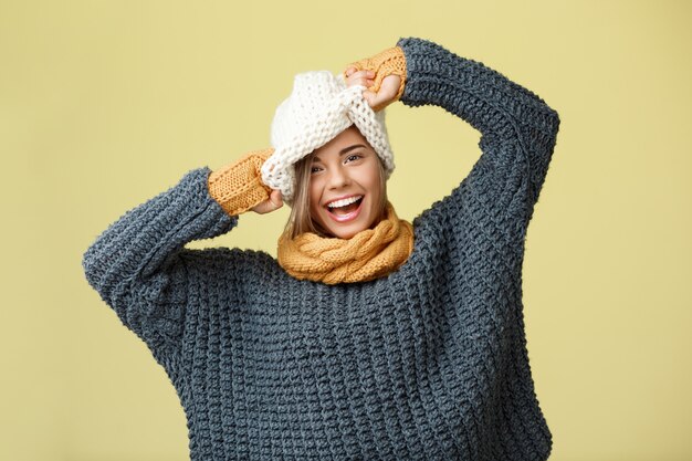 Young funny beautiful fair-haired woman in knited hat sweater scarf and mittens smiling on yellow.