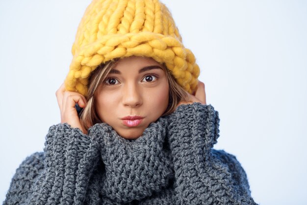 Young funny beautiful fair-haired woman in knited hat and sweater smiling on blue.