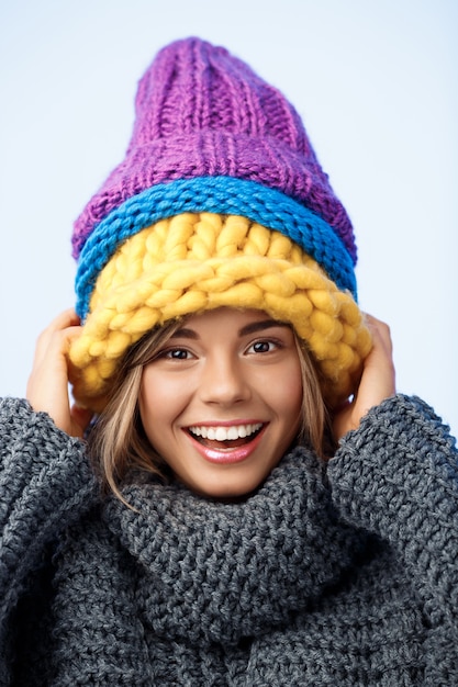 Young funny beautiful fair-haired woman in knited hats and sweater smiling on blue.