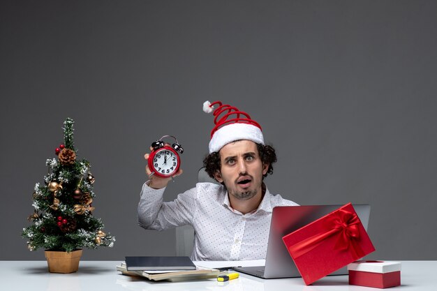 Young funny emotional excited business person with santa claus hat and holding and showing clock sitting in the office on dark background