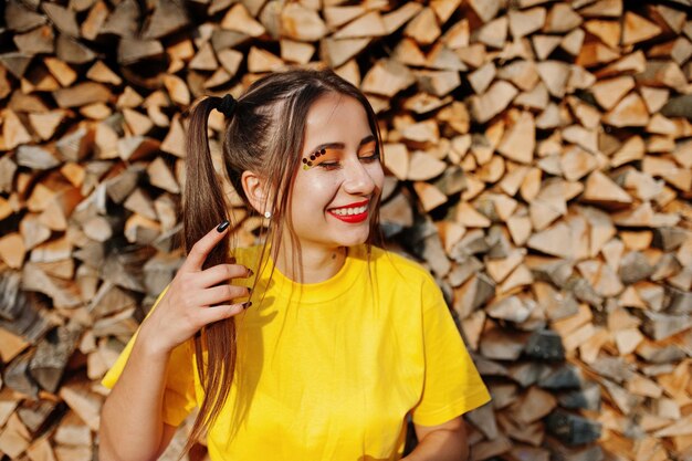 木製の背景に対して黄色のシャツに明るい化粧の尾の髪の若い面白い女の子