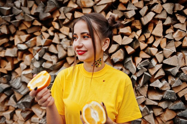 Молодая забавная девушка с ярким макияжем в желтой рубашке держит кусочек апельсина на деревянном фоне