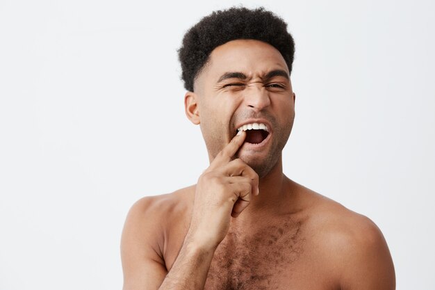 Young funny good-looking afro american male with curly hair without clothes picking in teeth looking at mirror with mean expression taking bath in morning.