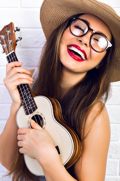 Young funny hipster woman having fun and playing on small ukulele guitar, singing and dancing. wearing vintage glasses and straw hat, joy, positive mood. 