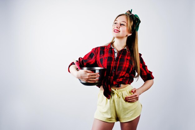 Young funny housewife in checkered shirt and yellow shorts pin up style with saucepan and kitchen spoon isolated on white background
