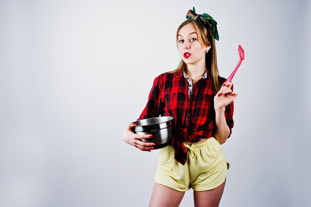 Young funny housewife in checkered shirt and yellow shorts pin up style with saucepan and kitchen spoon isolated on white background