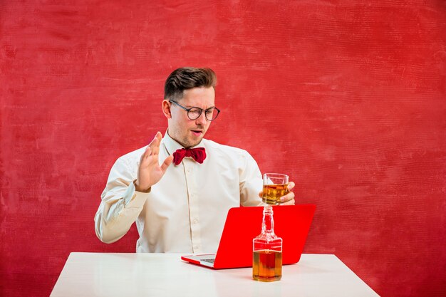 The young funny man with cognac sitting with laptop at St. Valentine's Day on red studio background. Concept - unhappy love