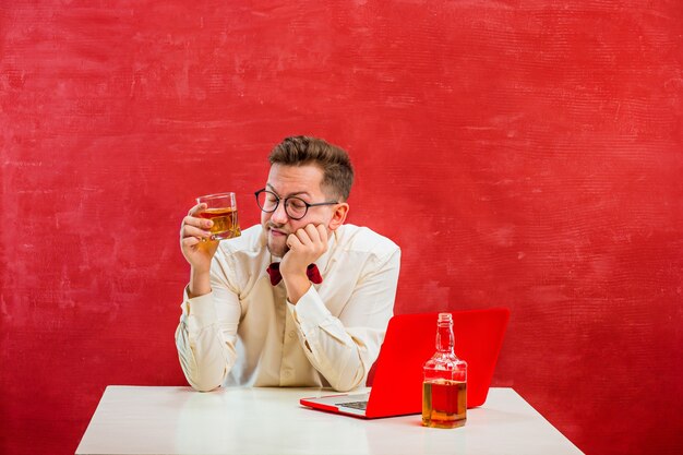 The young funny man with cognac sitting with laptop at St. Valentine's Day on red studio background. Concept - unhappy love