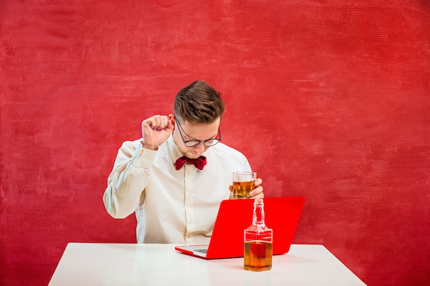 Giovane uomo divertente con cognac seduto con il computer portatile a san valentino su sfondo rosso studio.