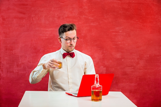 The young funny man with cognac sitting with laptop at st. valentine's day on red studio.