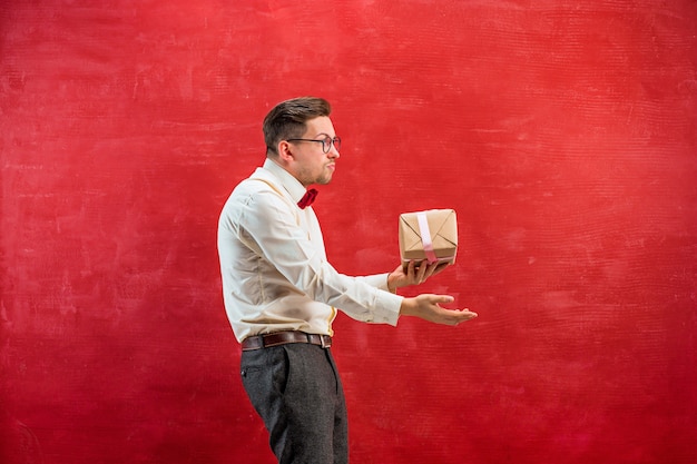 Young funny man with gift on red studio background