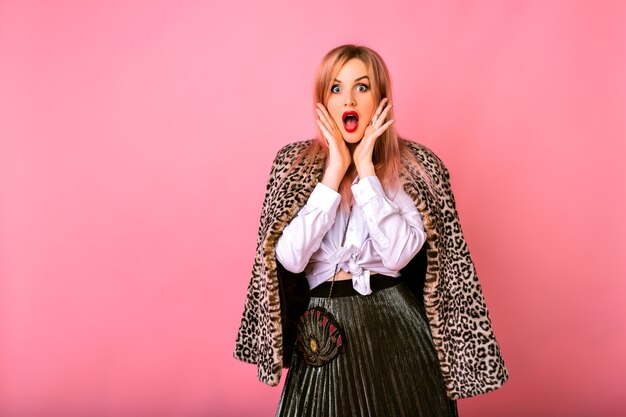 Young funny surprised shocked woman posing on pink background , wearing white shirt and leopard coat, powerful emotions.