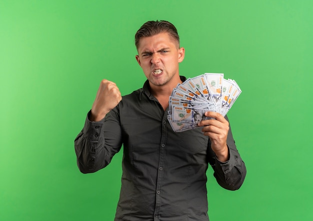 Free photo young furious blonde handsome man holds money with raised fist isolated on green space with copy space