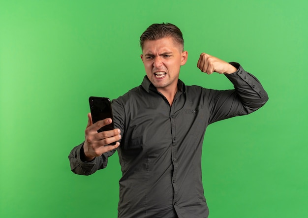 young furious blonde handsome man looks at phone and keeps fist up ready to punch