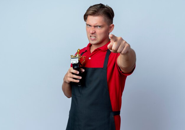 Young furious blonde male barber in uniform holds winner cup and hair clipper pointing at camera isolated on white space with copy space