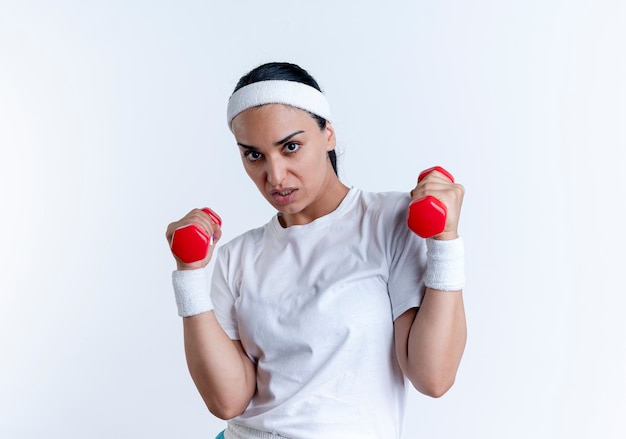 Young furious caucasian sporty woman wearing headband and wristbands holds dumbbells isolated on white space with copy space