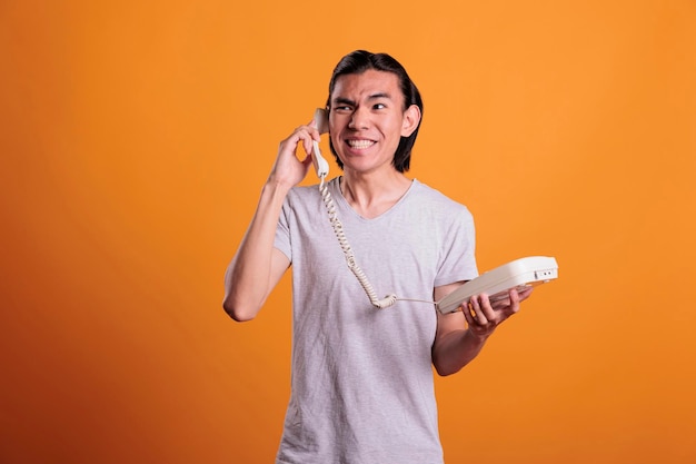 Young furious man talking on landline phone, shouting. Angry asian teenager screaming, holding retro telephone, person with aggressive facial expression standing, having conversation