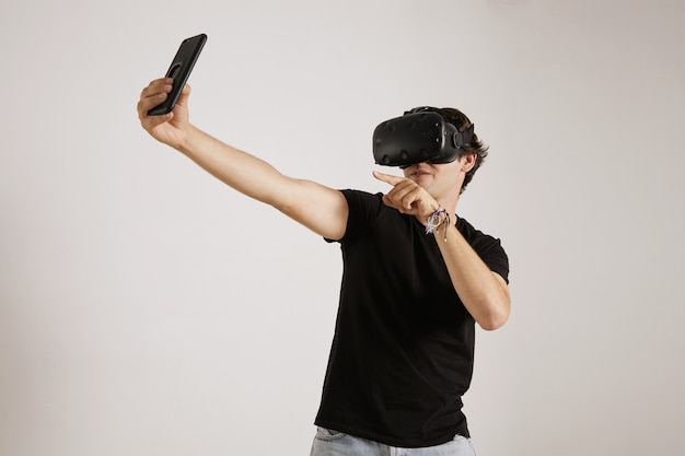 Молодой геймер в черной футболке и гарнитуре VR позирует для селфи на своем смартфоне на белой стене