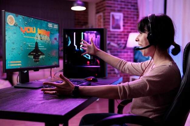 비디오 게임 챔피언십 우승을 축하하는 젊은 게이머는 온라인 라이브 스트림에서 네온 불빛이 있는 책상에서 행복함을 느낍니다. 컴퓨터에서 여성 스트리머 우승 사수 게임 토너먼트.