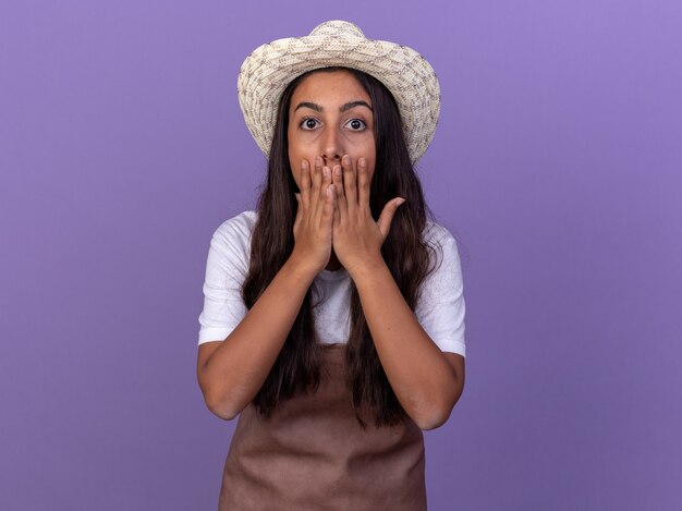 エプロンと夏の帽子をかぶった若い庭師の女の子は、紫色の壁の上に立っている手で口を覆って驚いてショックを受けました