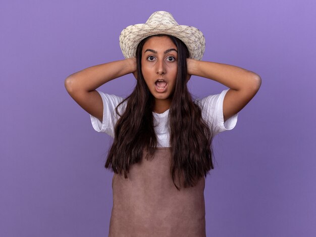 Young gardener girl in apron and summer hat  amazed and surprised standing over purple wall