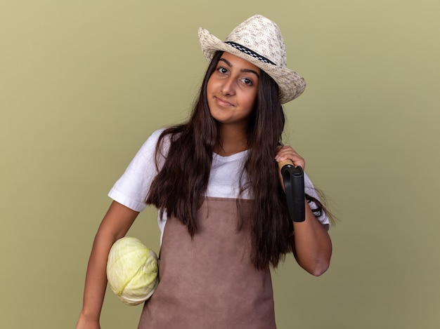 Young gardener girl in apron and summer hat holding cabbage and shovel  with smile on face standing over green wall