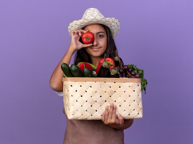 Молодая девушка-садовник в фартуке и летней шляпе держит ящик, полный овощей, прикрывая глаза свежими помидорами, стоящими над фиолетовой стеной