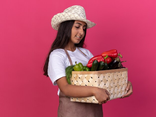 Young gardener girl in apron and summer hat holding crate full of vegetables looking confident smiling standing over pink wall